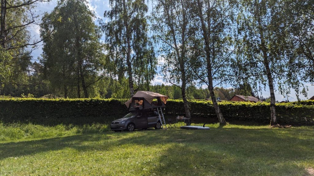 Auto mit aufgebautem Dachzelt auf einer Wiese am Waldrand, daneben Campingtisch und Stand-up-Paddle-Board